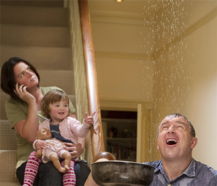 A man holding a pan catching water falling from the ceiling with a woman holding a child on the phone. 